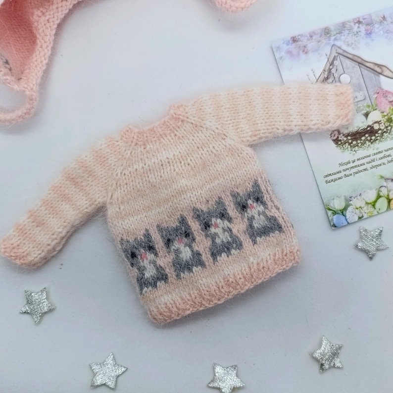 Puede incluir: Un su&eacute;ter tejido de color rosa claro con una fila de cuatro motivos de gatos grises y blancos en la parte delantera. La prenda de manga larga tiene un cuello redondo y dobladillo acanalados, confeccionada con hilo suave y textura sutil.