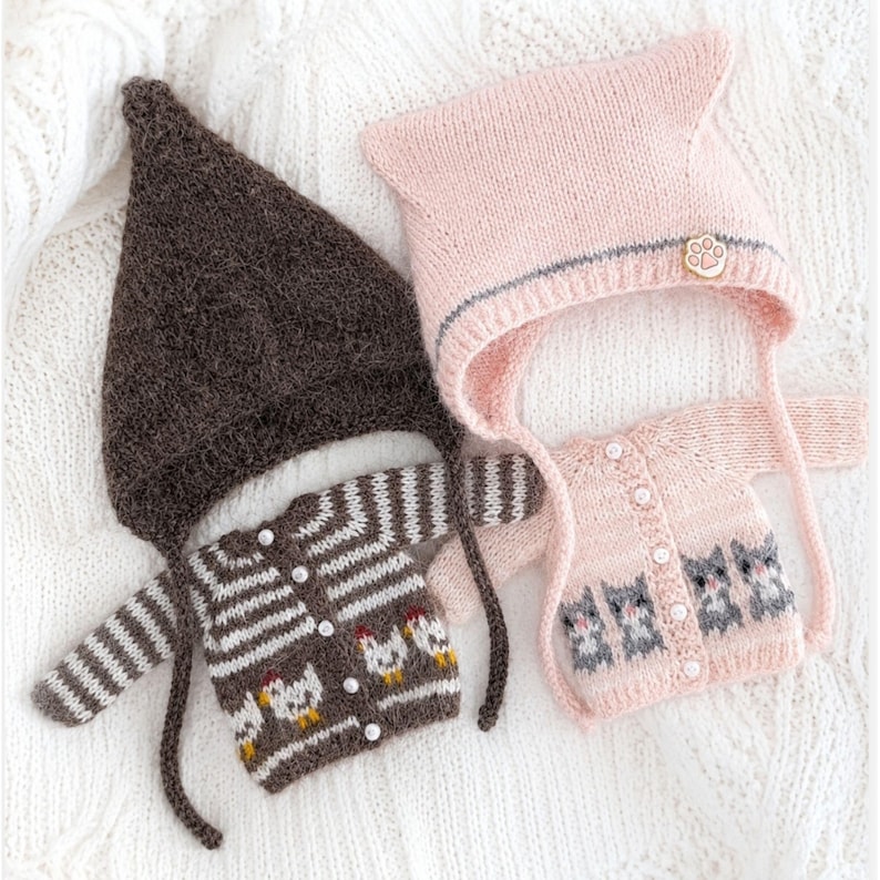 Puede incluir: Un gorro de punto marr&oacute;n oscuro en punta con una rebeca de rayas a juego con motivos de pollos blancos y botones nacarados, junto a un gorro de punto rosa claro con un bot&oacute;n de huella y una rebeca a juego con motivos de gatos grises.