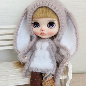 May include: A doll with large blue eyes and blonde hair, wearing a light gray bunny-eared hood and sweater. The sweater has a white bunny design on the front. The doll is wearing brown pants with a plaid patch.