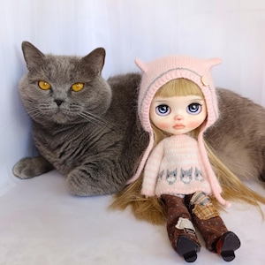 以下が含まれることがあります： 黄色い目をした灰色の猫が、長い金髪の人形の隣に座っています。人形は小さな耳が付いた薄いピンクのニット帽、猫の模様が入ったお揃いのピンクのセーター、パッチワークが施された茶色の柄入りパンツを着用しています。