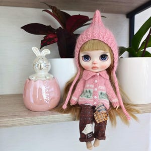 May include: A Blythe doll with long blonde hair, wearing a pink knitted hat and sweater with a green and white pattern. The doll is seated next to a pink ceramic Easter egg with a white bunny figurine inside.