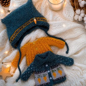 Pode incluir: Conjunto de roupa de bebé tricotada à mão. Um gorro azul-petróleo com abas para as orelhas e uma aplicação de cenoura, juntamente com dois camisolas em mostarda e azul-petróleo com desenhos de cenoura. Os artigos são apresentados sobre um tecido branco texturizado.