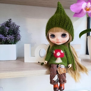Puede incluir: Una muñeca de pelo largo rubio que lleva un gorro de punto verde en punta y un jersey verde a juego con un gráfico de seta roja y blanca. La muñeca viste pantalones marrones estampados con un parche y botas marrones, sentada.