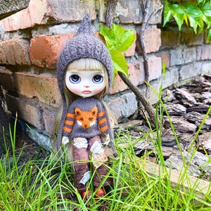 May include: A doll with blonde hair and large eyes wearing a grey knitted pointed hat, a grey sweater featuring an orange fox graphic with striped sleeves, and brown trousers with patches of various fabrics and patterns on the legs.