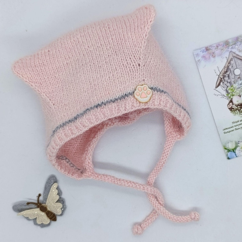 Puede incluir: Un gorro tejido de color rosa claro con una parte superior puntiaguda, una fina raya horizontal gris y un peque&ntilde;o emblema beige con forma de huella. El gorro incluye dos tiras tejidas y se muestra junto a un aplique decorativo de mariposa.