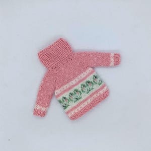 May include: A pink knitted turtleneck sweater featuring long sleeves with white cuffs and a horizontal band across the midsection decorated with a repeating green and white pattern. The garment is constructed with a ribbed collar and hem.