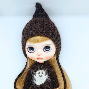 Pode incluir: Uma boneca com longos cabelos loiros usando um gorro de tricô pontudo marrom escuro e um suéter marrom escuro combinando com um gráfico de fantasma branco na frente. A boneca tem olhos azuis grandes, pele pálida e bochechas rosadas.