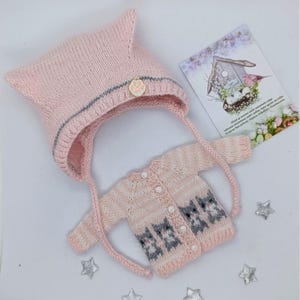 Puede incluir: Un gorro de punto rosa claro con una raya plateada y un dije de huella dorada, junto a un c&aacute;rdigan de manga larga a juego con un patr&oacute;n de gatos grises, botones blancos y una peque&ntilde;a tarjeta rectangular con un dibujo de pajarera.