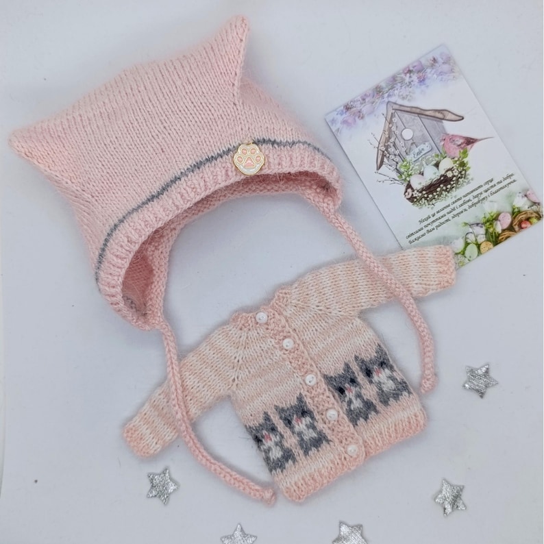 Puede incluir: Un gorro de punto rosa claro con una raya plateada y un dije de huella dorada, junto a un c&aacute;rdigan de manga larga a juego con un patr&oacute;n de gatos grises, botones blancos y una peque&ntilde;a tarjeta rectangular con un dibujo de pajarera.