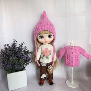 May include: A doll with long blonde hair wearing a pink cable-knit pointed hat, a white long-sleeved sweater with a pink flower design, and brown polka-dot pants with patches. Beside it, a pink cable-knit cardigan hangs on a white mannequin.