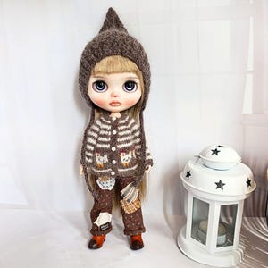 Puede incluir: Una mu&ntilde;eca de cabello rubio que viste un gorro puntiagudo de punto marr&oacute;n, un c&aacute;rdigan a rayas marrones y blancas con motivos de pollitos y botones blancos, y pantalones marrones estampados con parches, junto a un farol blanco con estrellas.