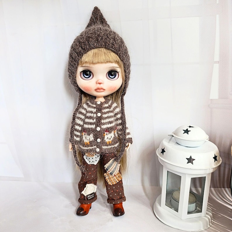 Puede incluir: Una mu&ntilde;eca de cabello rubio que viste un gorro puntiagudo de punto marr&oacute;n, un c&aacute;rdigan a rayas marrones y blancas con motivos de pollitos y botones blancos, y pantalones marrones estampados con parches, junto a un farol blanco con estrellas.