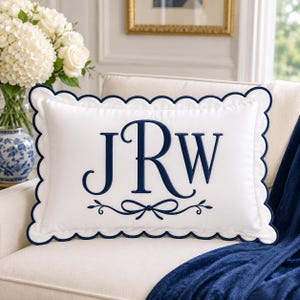 May include: A rectangular white throw pillow featuring a scalloped edge trimmed in navy blue. The center displays the embroidered navy blue monogram letters JRW, accented by a decorative bow and vine motif positioned directly below the lettering.