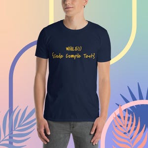 Könnte beinhalten: Marineblaues T-Shirt mit dem gelben Text "WHILE(1) {Code Compile Test}". Das Shirt hat kurze Ärmel und einen Rundhalsausschnitt. Der Hintergrund hat ein Pastellfarbschema.