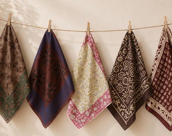 May include: Five square fabric handkerchiefs hanging from a horizontal string with wooden clips. Each cloth features a distinct pattern, including floral motifs, geometric shapes, and paisley designs in shades of brown, blue, pink, and cream.