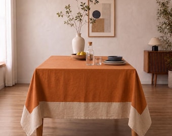 May include: A rectangular dining table covered with a burnt orange tablecloth featuring a wide beige border along the bottom edge. On the table sits a white ceramic vase with branches, a glass carafe, a small bowl with pears, and stacked plates.