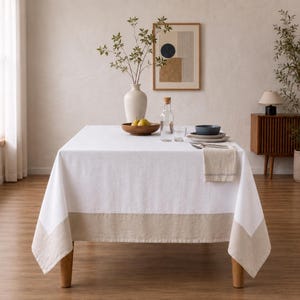 Puede incluir: Una mesa de comedor rectangular cubierta con un mantel blanco que presenta un borde ancho de color beige en la parte inferior. Sobre la mesa hay un jarr&oacute;n blanco con ramas verdes, un cuenco de madera con dos peras, una jarra y platos.
