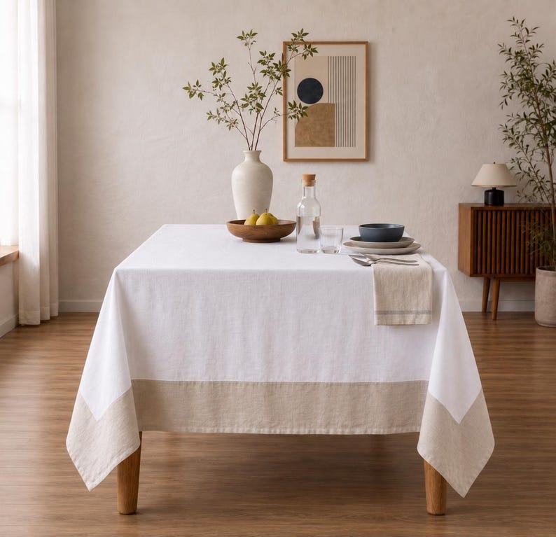 Puede incluir: Una mesa de comedor rectangular cubierta con un mantel blanco que presenta un borde ancho de color beige en la parte inferior. Sobre la mesa hay un jarr&oacute;n blanco con ramas verdes, un cuenco de madera con dos peras, una jarra y platos.