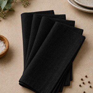 May include: A stack of four folded black cloth napkins. Each napkin features a square shape with visible fabric texture and straight, stitched edges. The napkins are layered neatly on top of one another, creating a uniform and structured appearance.