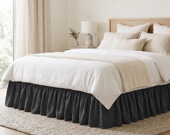 May include: A bed featuring a dark charcoal gray ruffled bed skirt made of fabric that drapes to the floor. The bed is dressed in white and cream-colored bedding, including a flat sheet, a duvet, and several pillows with a textured finish.