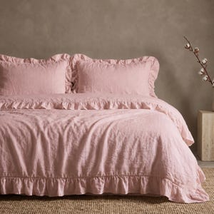 May include: A soft pink bedding set featuring a duvet cover and two matching pillowcases, each adorned with delicate ruffled edges. The fabric has a textured appearance, and the set is arranged neatly on a bed to showcase the ruffled detailing.