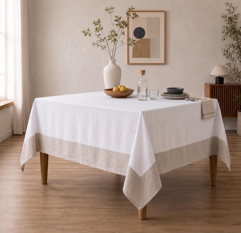 Puede incluir: Un mantel cuadrado blanco con un borde ancho de color beige sobre una mesa. Sobre la mesa hay un jarr&oacute;n blanco con ramas, un cuenco de madera con tres peras amarillas, una botella de cristal, un vaso y una pila de platos con una servilleta.