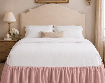 May include: A bed with a cream-colored headboard, white bedding, and a pink ruffled bed skirt. A small painting hangs above the bed. A nightstand with a lamp and flowers sits to the left. The room has a neutral color palette.