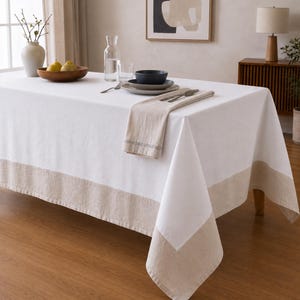 Puede incluir: Un mantel rectangular blanco con un borde ancho de color beige s&oacute;lido a lo largo del borde inferior. La tela tiene una textura suave y cae sobre los lados de una mesa, creando un aspecto limpio y neutro para el comedor.