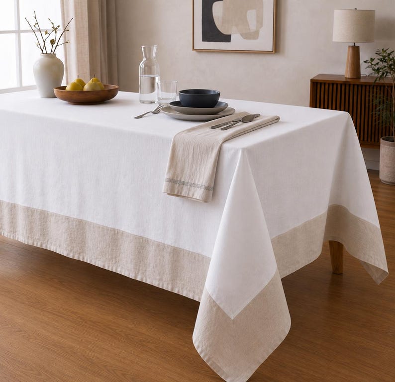 Puede incluir: Un mantel rectangular blanco con un borde ancho de color beige s&oacute;lido a lo largo del borde inferior. La tela tiene una textura suave y cae sobre los lados de una mesa, creando un aspecto limpio y neutro para el comedor.