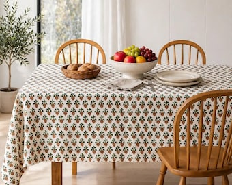 May include: A rectangular tablecloth featuring a repeating pattern of small brown pinecones with green needles on a white background. The fabric covers a wooden dining table surrounded by wooden spindle chairs, with a fruit bowl and bread basket.