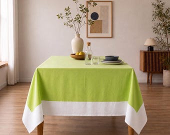 Puede incluir: Una mesa de comedor rectangular cubierta con un mantel de color verde lima sólido que presenta un borde blanco ancho a lo largo del borde inferior. La mesa está decorada con un jarrón de cerámica blanca con ramas, un cuenco de madera con peras y cristalería.