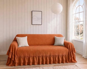 May include: A burnt orange sofa cover with a ruffled skirt along the bottom edge. The cover features a textured fabric and is accented by two square throw pillows, one white and one light gray, placed on the seat cushions.
