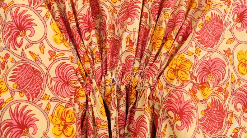 Puede incluir: Una prenda de tela de color amarillo claro con un patr&oacute;n repetitivo de motivos florales rosas y naranjas con detalles de enredaderas. El material est&aacute; fruncido en el centro con un cord&oacute;n blanco visible, creando pliegues.