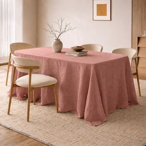 May include: A dusty rose linen tablecloth covers a rectangular table, set with a vase of branches, a bowl, and books. Four chairs with light wood frames and cream-colored seats surround the table, set on a neutral rug.