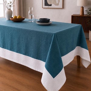 Puede incluir: Un mantel rectangular que presenta un panel central de color azul cerceta profundo bordeado por un ancho dobladillo blanco en los extremos. La tela tiene una textura tejida visible y cae sobre los lados de la mesa, creando un aspecto limpio y estructurado.