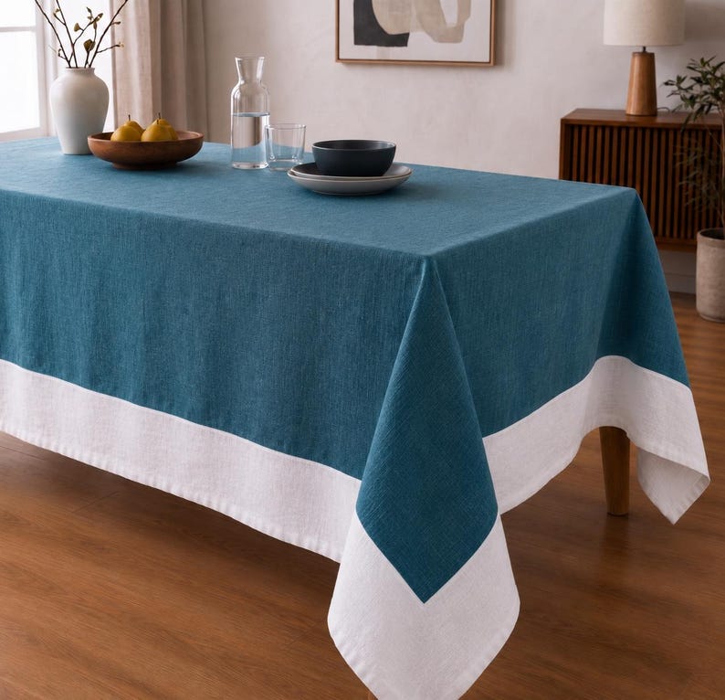 Puede incluir: Un mantel rectangular que presenta un panel central de color azul cerceta profundo bordeado por un ancho dobladillo blanco en los extremos. La tela tiene una textura tejida visible y cae sobre los lados de la mesa, creando un aspecto limpio y estructurado.