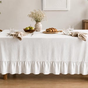 Pode incluir: Uma toalha de mesa retangular branca com uma borda franzida que cai da superfície da mesa. O tecido tem uma textura tecida visível e cobre toda a mesa, estendendo-se até o chão com pregas franzidas ao longo da bainha inferior.