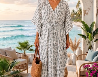 Op de afbeelding: Een enkellange witte kaftan-jurk met een herhalend grijs vispatroon, een V-hals en een taille met trekkoord. Het kledingstuk heeft wijde, losvallende mouwen en wordt gecombineerd met een gevlochten rieten tas die in één hand wordt gehouden.