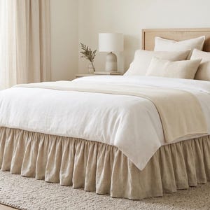 Könnte beinhalten: Ein Bett mit einem hellbeigen, gerüschten Bettvolant, einem weißen Bettbezug und einem passenden beigen Läufer. Das Set umfasst zwei große weiße Kissen, zwei beige Kissen und ein kleineres, beiges, rechteckiges Kissen vor einem Holzkopfteil.