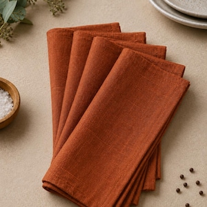 May include: A stack of four folded rectangular napkins in a solid burnt orange colour. The napkins feature a visible woven fabric texture with stitched edges, arranged in a slightly fanned overlapping pattern to display the textile weave.