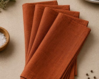 May include: A stack of four folded rectangular napkins in a solid burnt orange color. The napkins feature a visible woven fabric texture with stitched edges, arranged in a slightly fanned overlapping pattern to display the textile weave.