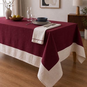 Puede incluir: Un mantel rectangular que presenta un panel central de color burdeos intenso y un borde ancho y contrastante de color crema a lo largo de los lados. La tela tiene una textura tejida visible y el mantel cubre una mesa con vajilla encima.