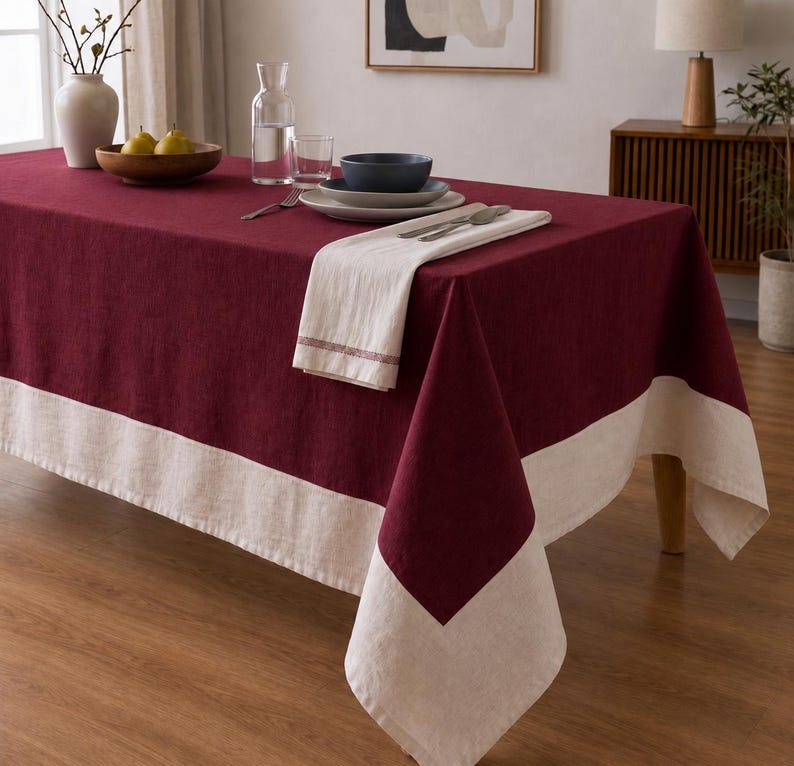 Puede incluir: Un mantel rectangular que presenta un panel central de color burdeos intenso y un borde ancho y contrastante de color crema a lo largo de los lados. La tela tiene una textura tejida visible y el mantel cubre una mesa con vajilla encima.