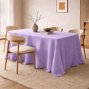 Puede incluir: Un mantel de lino lavanda cubre una mesa rectangular, con un jarrón de ramas, libros y un cuenco. Cuatro sillas de color claro rodean la mesa, sobre una alfombra de tonos neutros. Una imagen enmarcada cuelga de la pared.