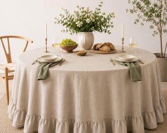 Puede incluir: Un mantel redondo de lino beige con borde con volantes. La mesa está puesta con platos, servilletas, cubiertos y copas de vino. Un jarrón de flores blancas y un cuenco de madera con uvas están en el centro.