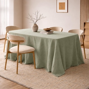 Puede incluir: Un mantel de lino verde salvia cubre una mesa de comedor rectangular. Cuatro sillas de color claro con respaldos curvos rodean la mesa. Un jarrón con ramas y un cuenco pequeño están sobre la mesa.