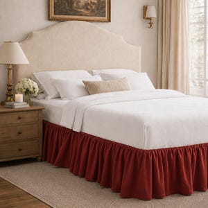 May include: A bedroom scene featuring a bed with a cream-colored headboard and white bedding. A deep red bed skirt adds a pop of color. A wooden nightstand with a lamp and a framed landscape painting complete the elegant decor.