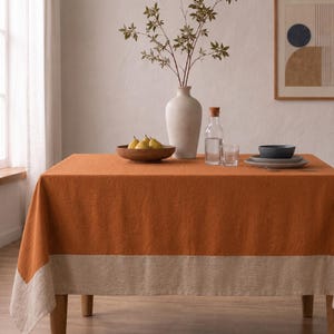 Puede incluir: Un mantel rectangular que presenta un panel central de color naranja quemado s&oacute;lido con un borde ancho y contrastante en color beige a lo largo del borde inferior. La tela tiene un tejido texturizado visible, drapeado sobre una mesa.