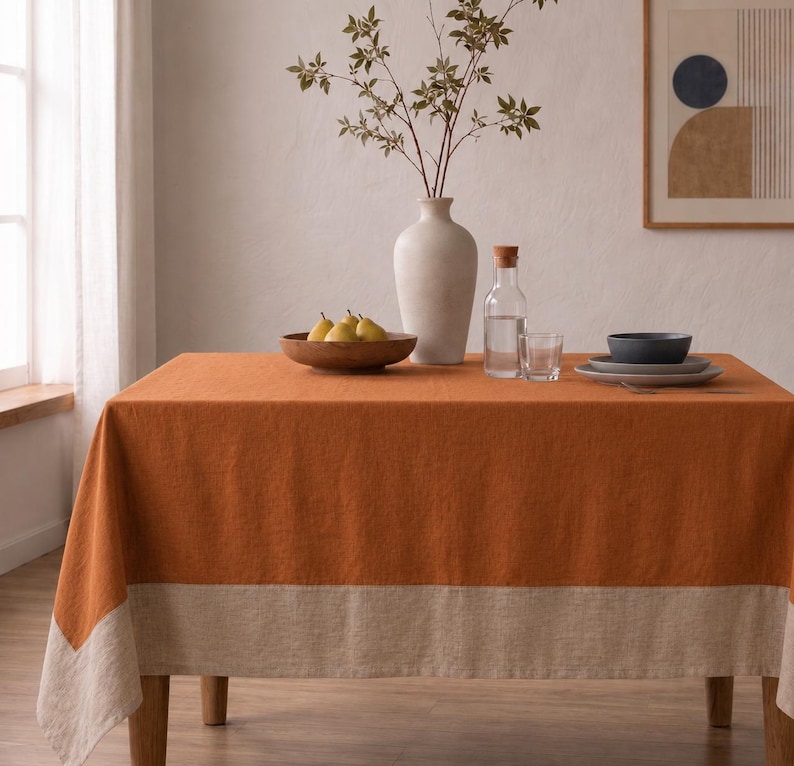 Puede incluir: Un mantel rectangular que presenta un panel central de color naranja quemado s&oacute;lido con un borde ancho y contrastante en color beige a lo largo del borde inferior. La tela tiene un tejido texturizado visible, drapeado sobre una mesa.