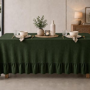 May include: A dark green rectangular tablecloth with a ruffled edge, draped over a dining table. The table is set with two white bowls on plates, gold-toned cutlery, clear glasses, a small potted plant, a candle, and a tall white vase.